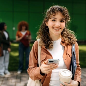 Person holding a smartphone and takeaway coffee cup outdoors, with people and a green building in background.