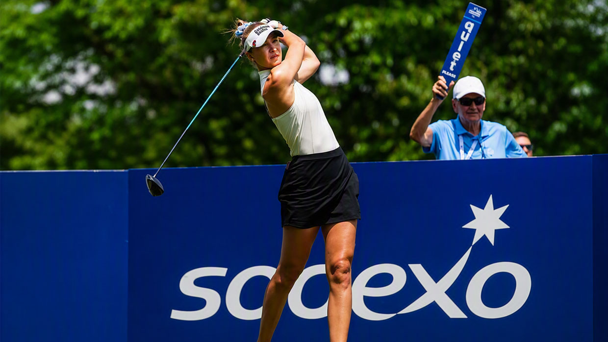 Golfer teeing off at a tournament with a blue sponsor board, trees, and course officials in the background.