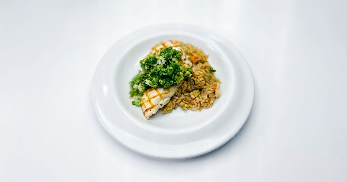 Grilled fish topped with green herb sauce served with seasoned vegetable rice on a white plate. 
