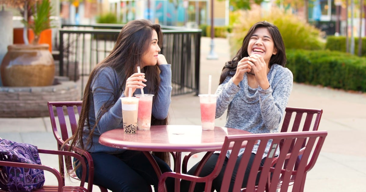 Two college students sit at an outdoor table laughing and talking, each with a boba drink in front of them.