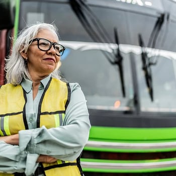 A bus driver in a safety vest stands confidently in front of a green bus.