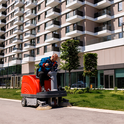 Commercial street sweeper cleaning a pedestrian walkway beside landscaped grounds and residential buildings