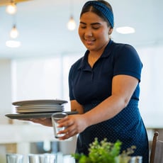 A young foodservice worker smiling while cleaning up a table in a restaurant.