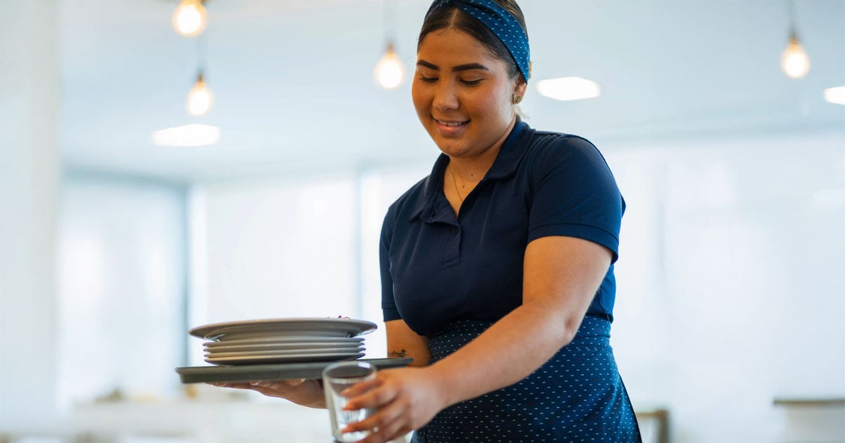 A young foodservice worker smiling while cleaning up a table in a restaurant.