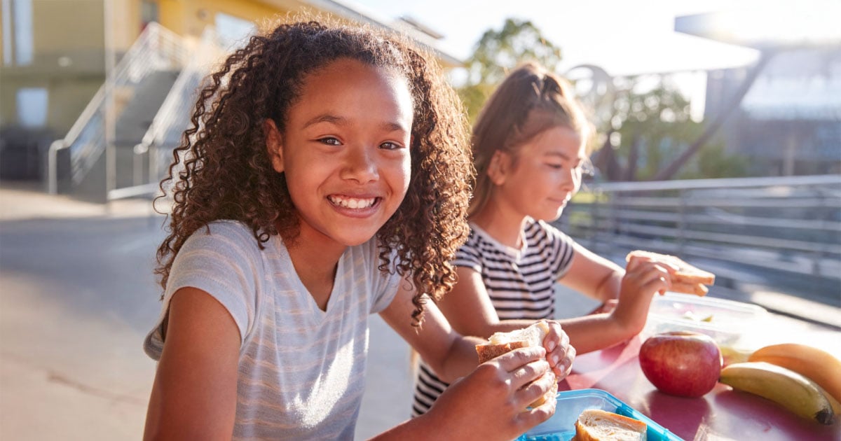 Two children eat sandwiches and fruit at a red outdoor table; one smiles at the camera.