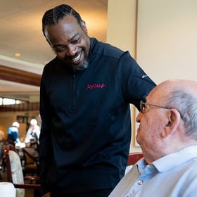Dining staff member warmly greeting a senior resident during mealtime.