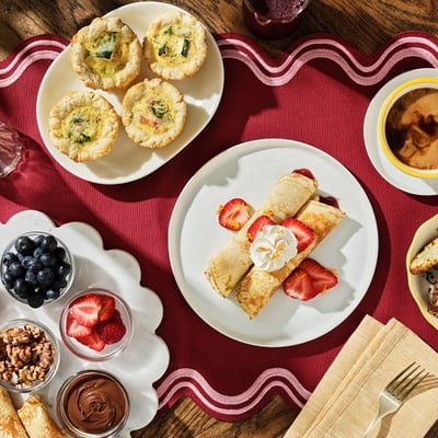 Overhead view of a brunch spread with crepes, mini quiches, fresh berries, and coffee on a stylish table setting. 