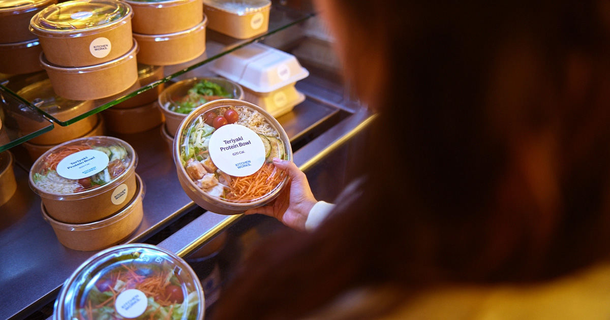 Person holding a Teriyaki Protein Bowl in front of a refrigerated shelf with other packaged meals.