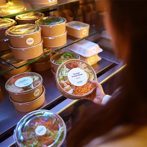 Person holding a Teriyaki Protein Bowl in front of a refrigerated shelf with other packaged meals.