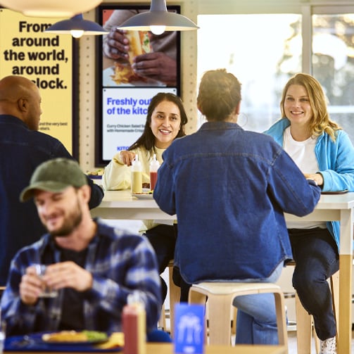 Four people sit at a table in a casual dining area with two posters visible in the background.