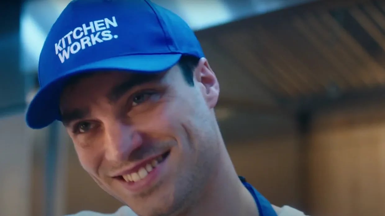 Person in a blue cap that says “KITCHEN WORKS.” stands in a kitchen, partially turned away from the camera.