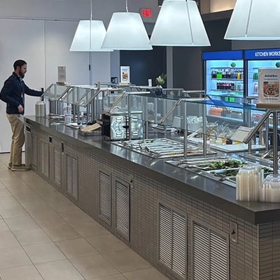 Man serving food at a salad bar in a modern cafeteria with drinks and food options behind.