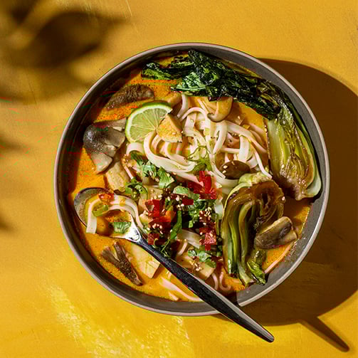 Bowl of noodle soup with bok choy, mushrooms, lime wedge, and red chilies in orange broth on yellow backdrop.
