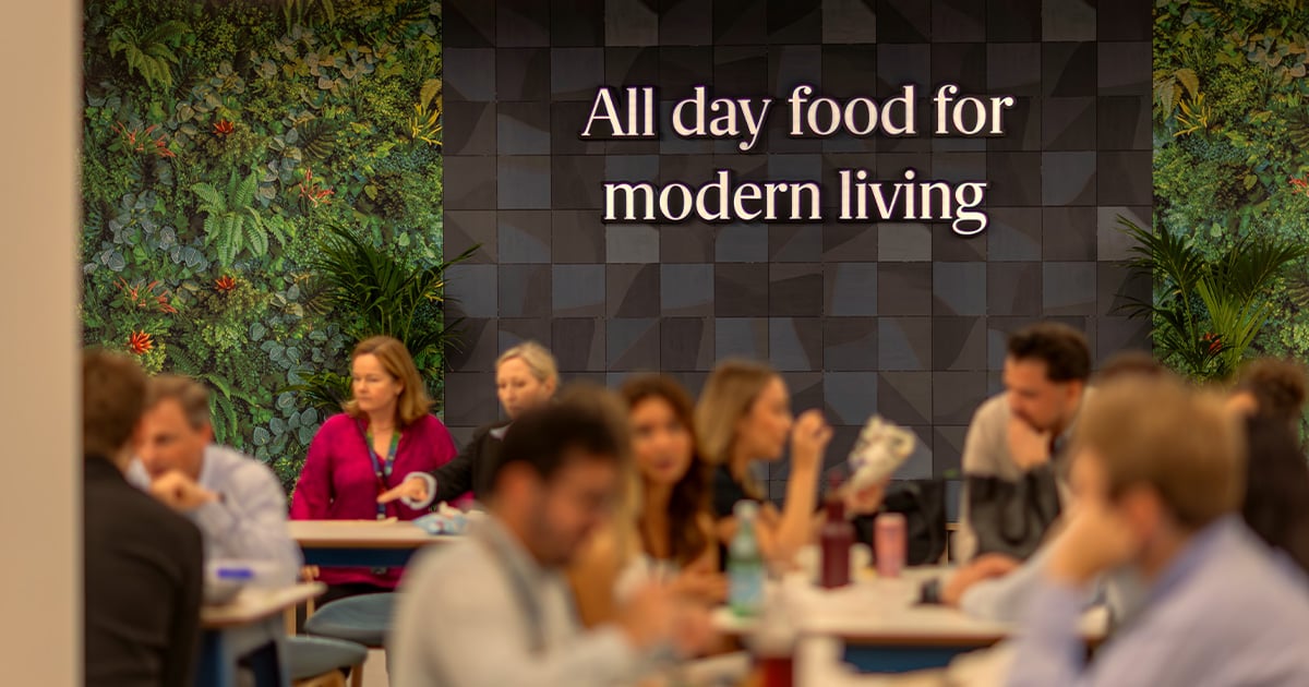 People sit in a cafeteria with a green plant wall and a sign reading “All day food for modern living.”