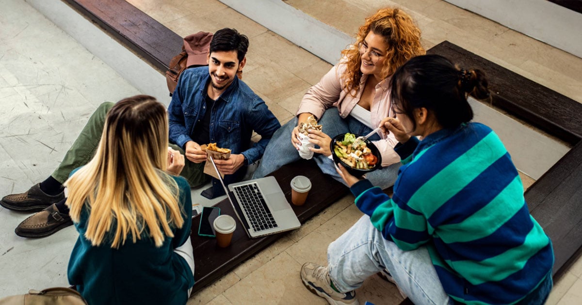 A group of college students sitting on courtyard steps, chatting and sharing lunch together.