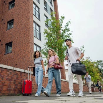 Group of young adults walking with luggage outside a campus-style brick building. 