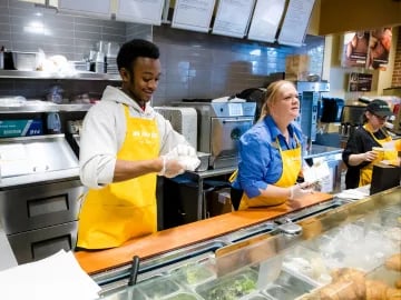 Three workers in yellow aprons behind a counter preparing food in a restaurant kitchen.