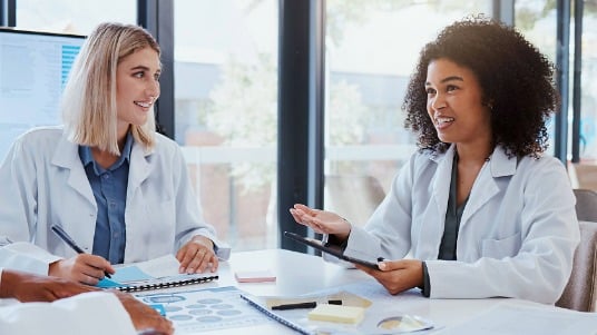 Two dietitians sit at a table reviewing research notes, with one holding a tablet and data displayed on a screen behind them.