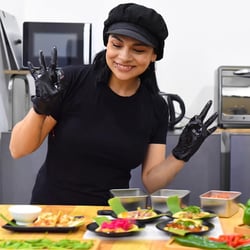Chef in black gloves and hat poses with peace signs behind a table of colorful food dishes.