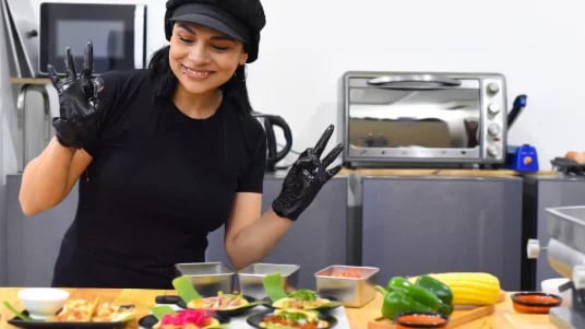 Chef in kitchen with black gloves, posing beside fresh vegetables and plated dishes on counter.