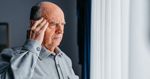Person standing by a bright window holding one hand to the side of the head.