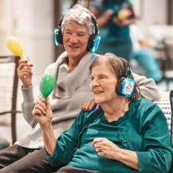 Photo Credit: The Day by Day Project Two senior residents wearing headphones and shaking maracas while enjoying music.