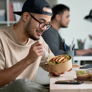 A male student wearing glasses and a backward cap eating a healthy meal at his desk. 