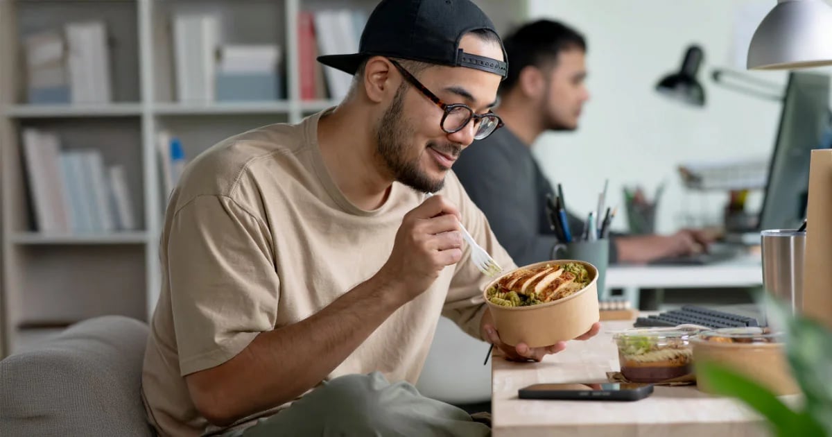 A male student wearing glasses and a backward cap eating a healthy meal at his desk. 
