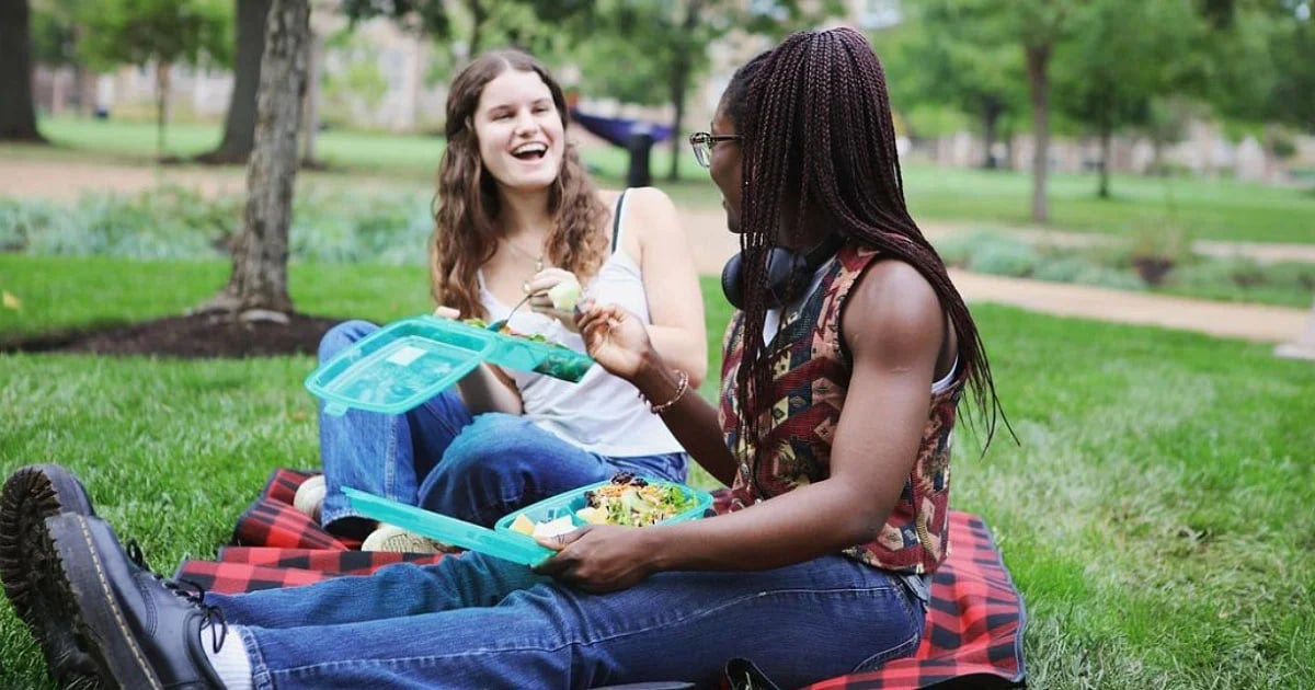 Two friends sit on a checkered blanket in a park, sharing a meal from reusable containers and smiling as they chat.