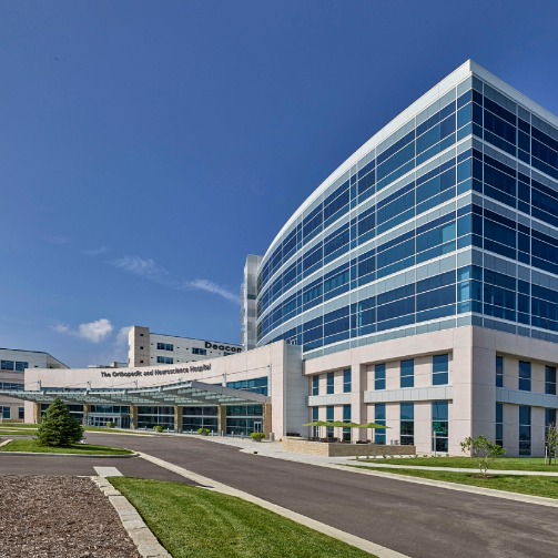 Modern hospital building with a curved glass façade, main entrance canopy, landscaped grounds, and access road, viewed under a clear blue sky during daylight.