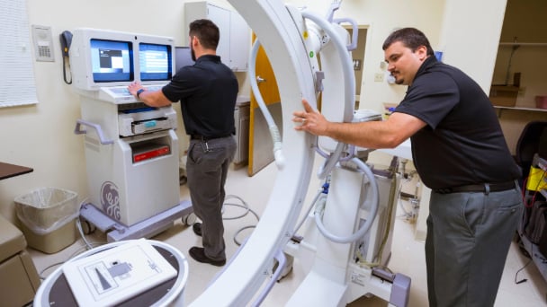 Two men operate a large machine in a hospital setting, focused on their task.