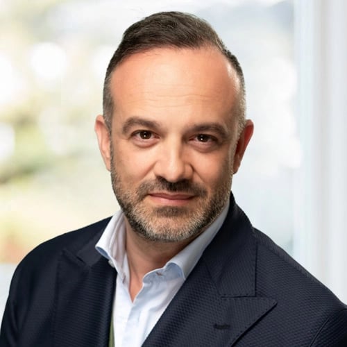 Person with short hair and beard in dark suit and white shirt, standing before a blurred background.