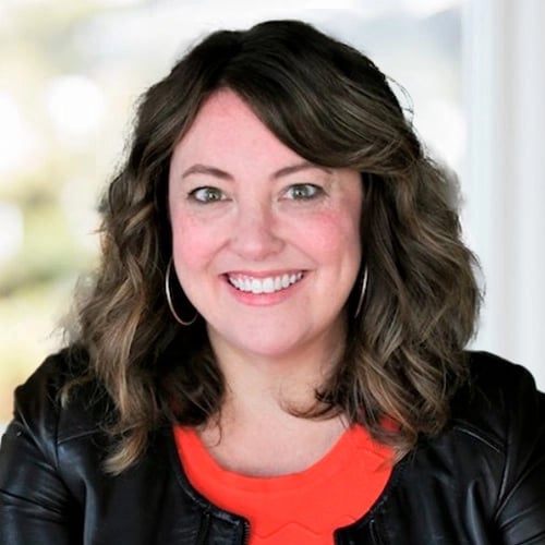 Person with shoulder-length wavy hair in black jacket and orange top, smiling at the camera.