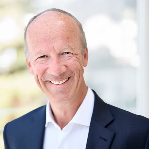 Person wearing a dark suit jacket and white collared shirt in a bright indoor setting.