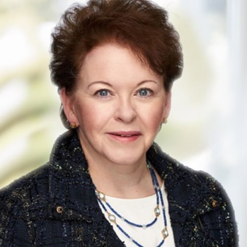 Studio headshot of an adult with short brown hair, wearing a dark blazer and layered necklace against a soft blurred background.