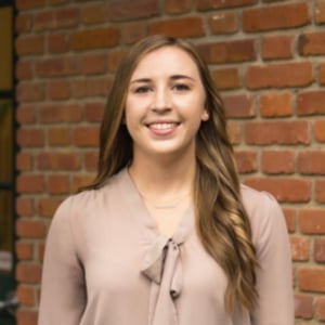 Person with long hair in light blouse stands in front of brick wall.