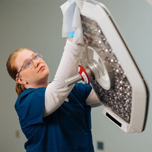 Healthcare environmental services professional cleaning and disinfecting an overhead surgical light in a clinical setting, highlighting infection prevention, hospital sanitation, equipment maintenance, and patient safety.