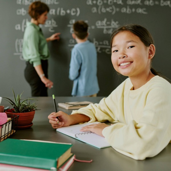 A student sitting at a desk in a classroom while a teacher and another student work on math equations at the chalkboard in the background.