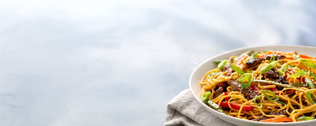 Close-up of an Asian-inspired beef stir-fry noodle bowl with vegetables and sprinkled sesame seeds. 