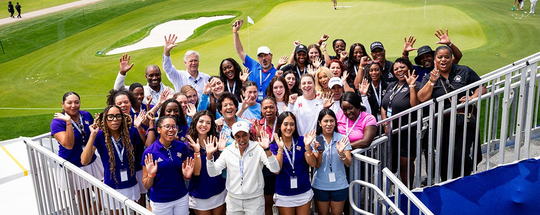 Group of event attendees waving on stairs beside a golf course green and practice area.
