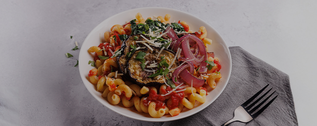 A bowl of pasta topped with grilled eggplant, red onions, spinach, and grated cheese.