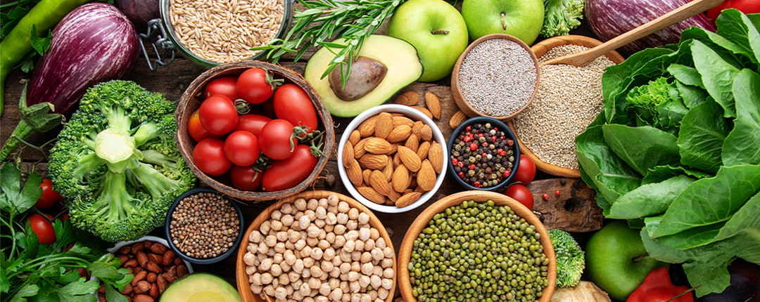 Top-down view of fresh whole foods including vegetables, fruits, grains, nuts, and legumes arranged in bowls on a wooden surface, highlighting healthy and diverse ingredients.