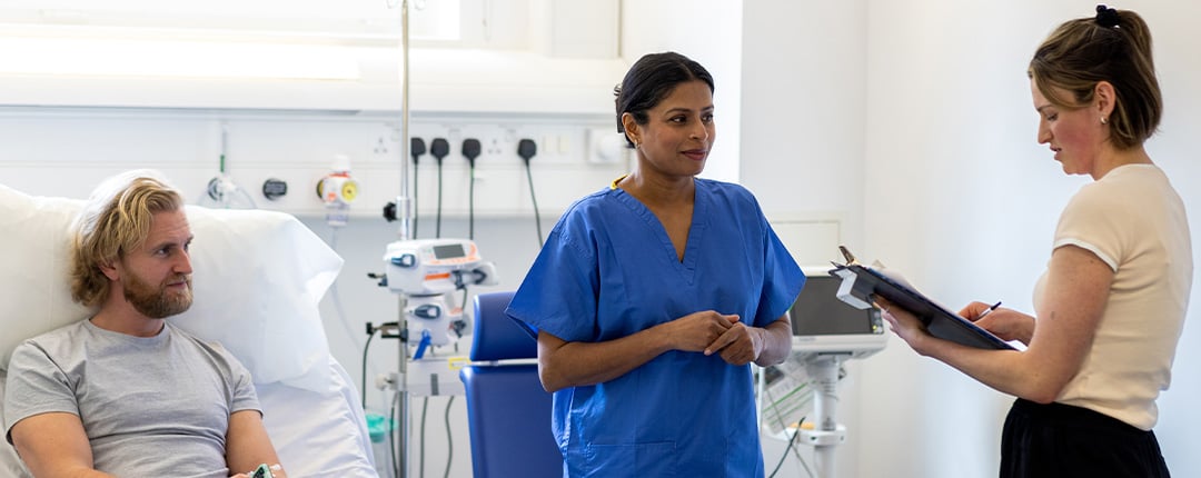 Healthcare professionals reviewing patient care information at a hospital bedside, with medical equipment visible in a bright clinical environment focused on care coordination and treatment planning.