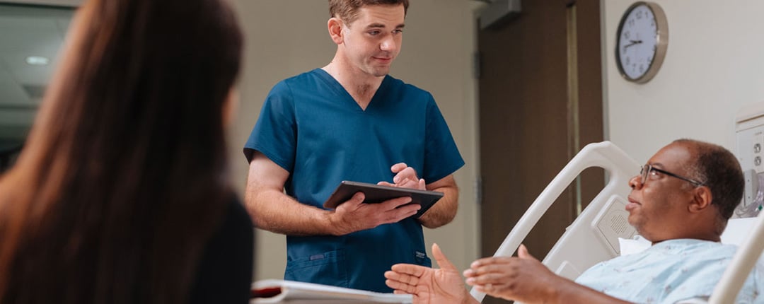 Healthcare professional in blue scrubs holding a tablet while speaking with a patient resting in a hospital bed, with another person seated nearby and medical room furnishings visible in the background.