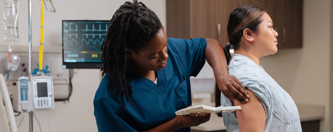 Healthcare professional in scrubs measuring a patient’s upper arm with a body composition assessment device in a hospital room, with medical equipment and monitoring screens visible in the background.