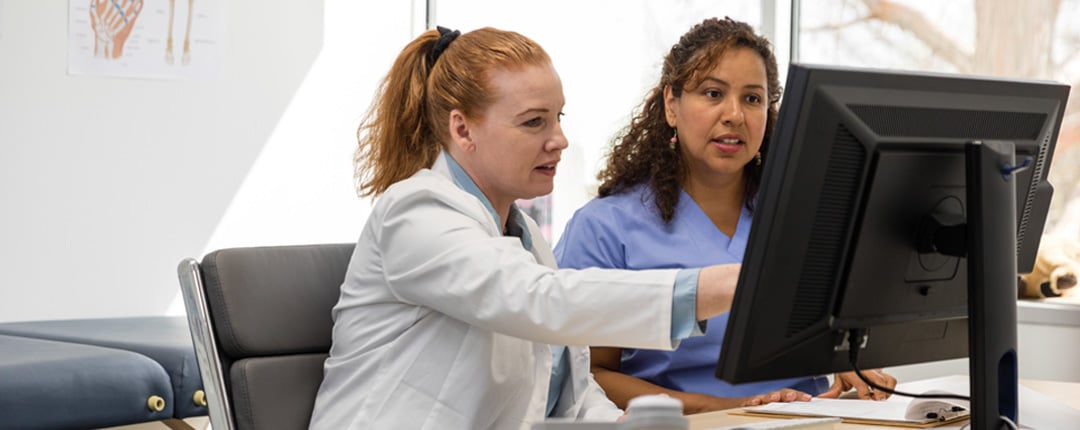 Two healthcare professionals sit at a desk in a bright medical office, looking at a desktop computer monitor.