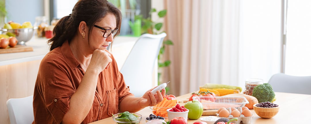 Person reviewing a glucose meter at a kitchen table surrounded by fresh vegetables, eggs, berries, and healthy ingredients, highlighting diabetes management, nutrition planning, and informed dietary choices.