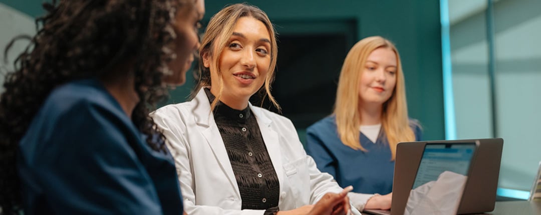 Three people in medical attire are seated around a desk, with one person in a white coat actively leading the discussion and gesturing, while another in scrubs listens from the side.