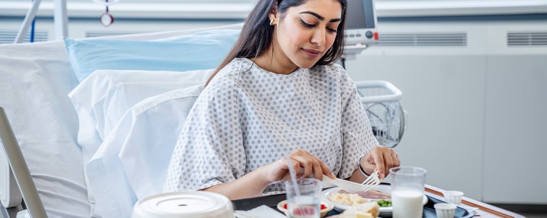 Hospital patient seated in bed eating a meal from a tray, with medical equipment and bedding visible.
