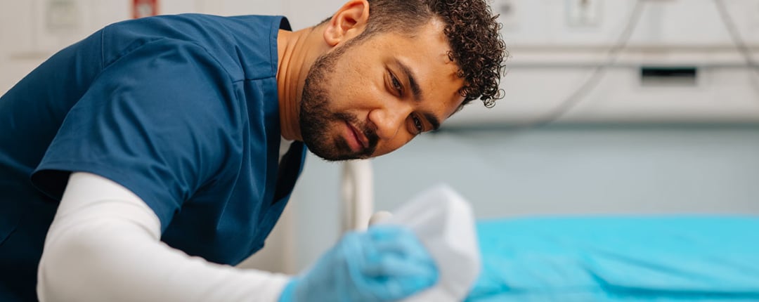Environmental services worker cleaning and disinfecting a hospital bed with a cloth, highlighting healthcare sanitation, infection prevention, patient safety, and facility hygiene practices.
